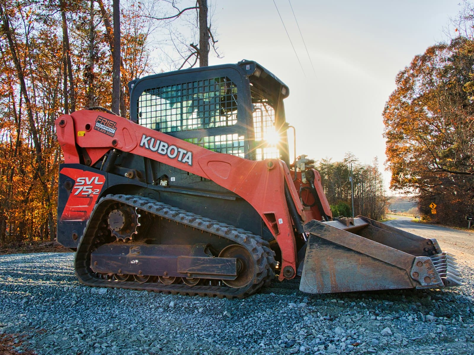 A compact track loader standing on a gravel yard at the edge of an Ozark hardwood stand.